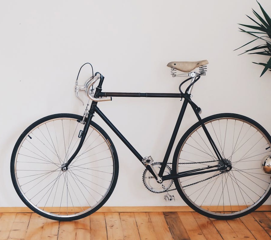 Black vintage road bicycle with a tan leather saddle leaning against a white wall on a light wooden floor